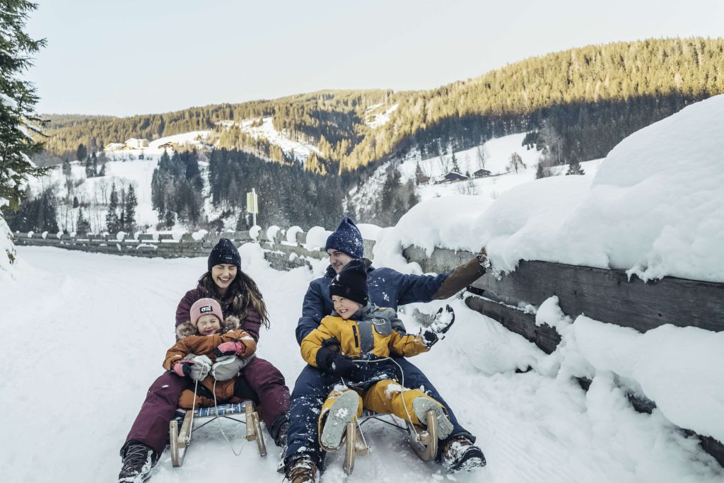 Familie vergnügt sich beim Schlittenfahren im verschneiten Kaprun vor malerischer Bergkulisse.
