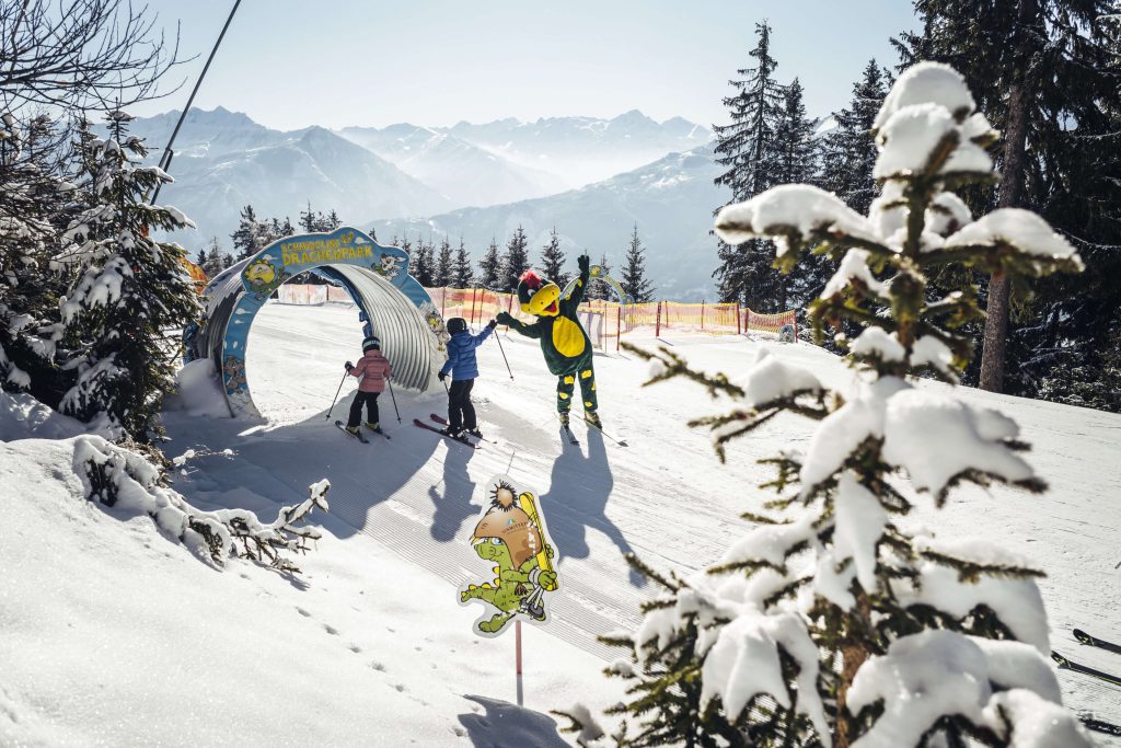 Kinder lernen Skifahren im Spaßpark mit schneebedeckten Bergen im Hintergrund.